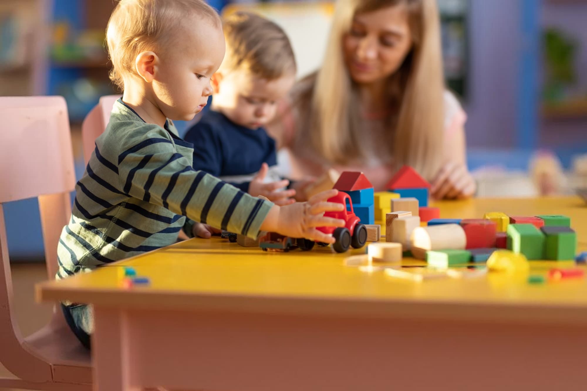 A toddler playing with a toy truck and colorful wooden blocks at a classroom table, with another child and a teacher behind him