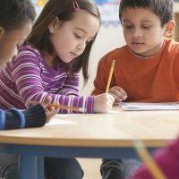 Three children at a classroom table with paper and pencil.